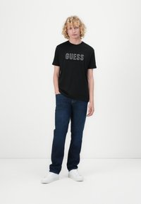 Svart bomull t-skjorte med "GUESS" logo i hvitt, kombinert med mørkeblå denimjeans og hvite sneakers. Enkel, uformell design.