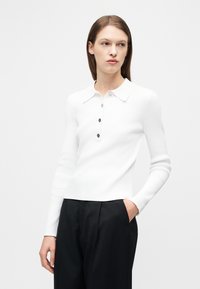 Γυναίκα που φοράει λευκό ριμπ πικέ πόλο με μακρύ μανίκι και μαύρα κουμπιά, σε συνδυασμό με μαύρο ψηλόμεσο παντελόνι, όρθια με το ένα χέρι στην τσέπη.