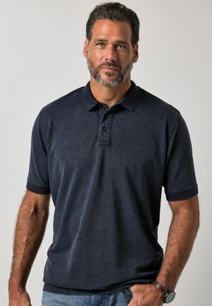 Mand med kort mørkt hår og skæg iført en mørkeblå poloshirt og jeans, stående mod en ensartet lys baggrund.