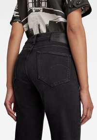 Personne portant un jean noir taille haute avec poches arrière et une chemise à manches courtes à motif noir et blanc.