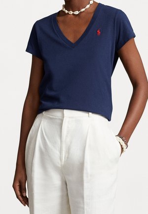 Woman wearing a navy blue V-neck shirt with red logo, white high-waisted pants, shell necklace, and bracelets on left wrist.