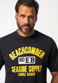Námořnické tričko s výraznou žlutou grafikou, na které je napsáno "BEACHCOMBER", "80 EB" a "SEASIDE SUPPLY". Vyrobeno z měkké tkaniny s kulatým výstřihem.