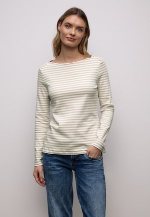 Jeune femme aux cheveux châtain clair portant un chemisier à rayures beige et blanc à manches longues et un jean bleu, debout avec une main dans la poche.