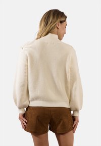 Maglione crema a coste con colletto alto, maniche a palloncino e lavorazione a maglia testurizzata, abbinato a pantaloni corti in suede marrone.