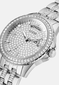 Srebrny zegarek Guess z krystaliczną obręczą i tarczą, z wyświetlaczem daty i wskaźnikiem dnia, na stalowej bransolecie.