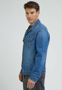 Mężczyzna w niebieskiej dżinsowej kurtce i ciemnych spodniach, stojący bokiem i patrzący w stronę aparatu na tle jasnego, jednolitego tła.