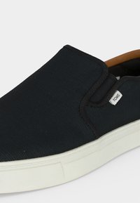 Chaussure noire en toile à enfiler avec semelle en caoutchouc blanche et petite étiquette en tissu marquée TOMS sur le côté près de l'ouverture.