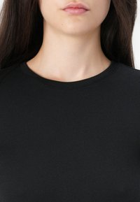 Giovane donna con lunghi capelli scuri, che indossa una maglietta nera a girocollo semplice, visibile dalla parte bassa del viso fino alla parte alta del petto.