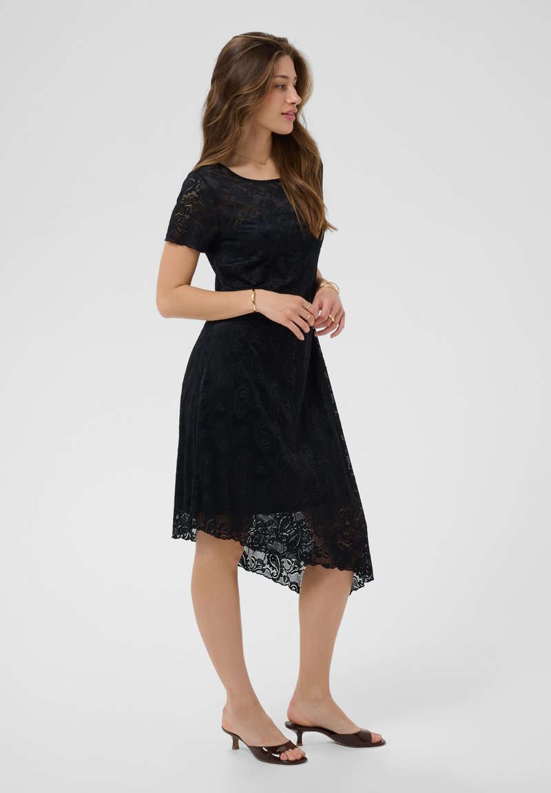 Femme aux longs cheveux portant une robe courte en dentelle noire à manches courtes et des escarpins ouverts marron, debout de profil devant un fond uni.
