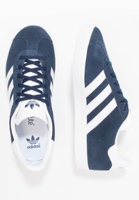 Marina mockasiner i mocka med vita gummisulor, med tre vita ränder på varje sida och svart adidas-logotyp på tungan.