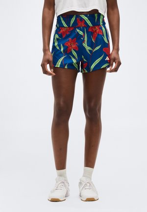 Inimene, kes kannab siniseid pükse punase lillemustriga, valgeid pahkluu sokke ja valgeid spordijalatsid ühetaolise tausta ees.