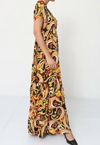 Robe maxi noire avec un motif paisley jaune, rouge et orange. Manches courtes, corsage ajusté et jupe évasée avec volants en niveaux.