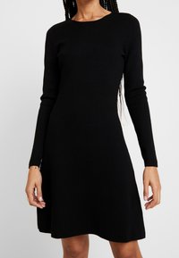 Schwarzes Strickkleid mit langen Ärmeln, rundem Halsausschnitt und schwingendem Rock. Weicher Stoff und kniebedeckende Länge, minimalistisches Design ohne Muster.