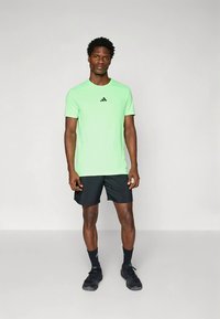 Světlé zelené atletické tričko s kulatým výstřihem, černé logo Adidas na hrudi, doplněné černými šortkami a atletickou obuví.