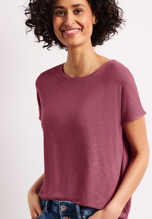 Mujer con cabello rizado sonriendo, lleva una camiseta de manga corta, cuello redondo color malva y vaqueros azules con las manos en los bolsillos.