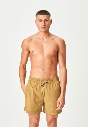 Muž s výraznou svalnatou postavou a krátkymi vlasmi, oblečený v hnedých plavkách, stojaci s rukami voľne pozdĺž tela na jednoduchom svetlom pozadí.