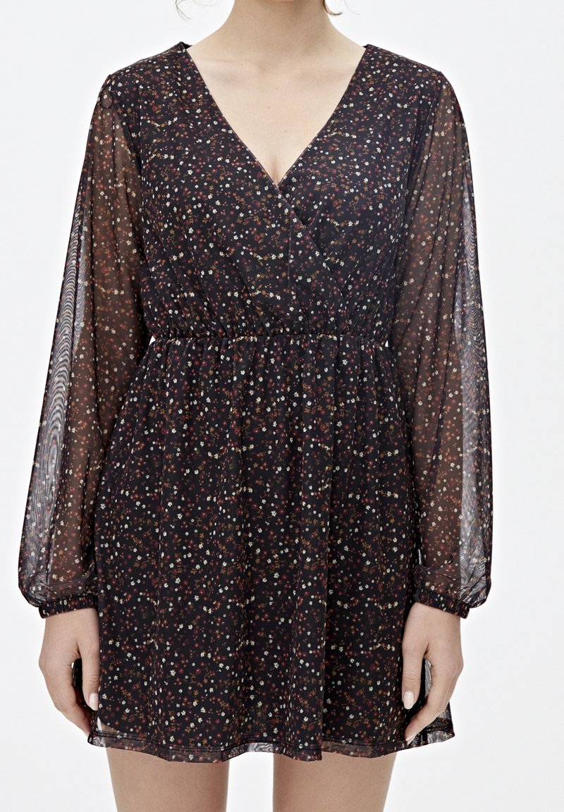 Robe longue noire à manches longues avec petit imprimé floral multicolore, col en V, taille froncée et manches transparentes sur une personne vue du cou jusqu'à la moitié des cuisses.