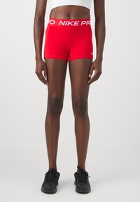 Red Nike Pro shorts with a white logo waistband, featuring a fitted design and smooth texture. Worn with black athletic shoes.