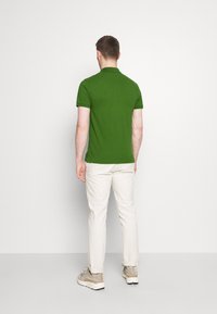 Homme debout, de dos, portant un polo vert à manches courtes, un pantalon blanc et des baskets beiges sur un fond uni.