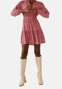 Rotes Blumenmuster-Kleid mit gerafftem Oberteil, langen Ärmeln und gestuftem Rock, kombiniert mit beigen kniehohen Stiefeln. Glatte Textur, Knopfdetails.