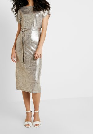 Donna che indossa un vestito argento lucido al ginocchio con maniche corte e una cintura abbinata, abbinato a sandali bianchi con tacco alto.