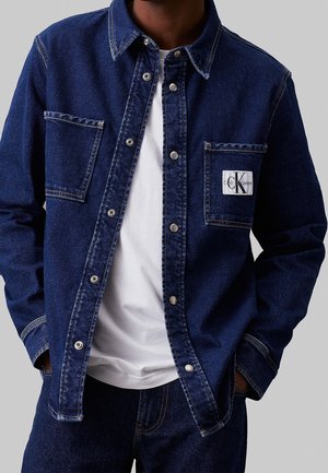 Osoba nosząca ciemnoniebieską kurtkę jeansową z zatrzaskami i dwoma przednimi kieszeniami, z których jedna posiada naszywkę z logo Calvin Klein Jeans.