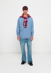 Uomo che indossa un maglione azzurro chiaro, una camicia a quadri rossi annodata intorno alle spalle, jeans blu, scarpe nere e un berretto bordeaux, in piedi contro uno sfondo bianco.