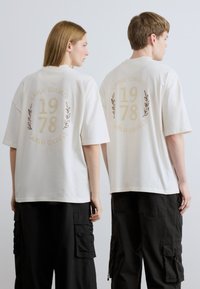 Λευκά υπερμεγέθη t-shirts με επιγραφή "CARLO COLUCCI 1978" σε χρυσή εκτύπωση, διακοσμημένα με φυλλαράκια δάφνης στην πλάτη. Λεπτομέρειες από απαλό βαμβακερό υλικό.