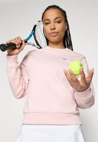 Kvinne iført lys rosa hettegenser og hvit skjørt som holder en tennisracket over skulderen og kaster en gul tennisball.