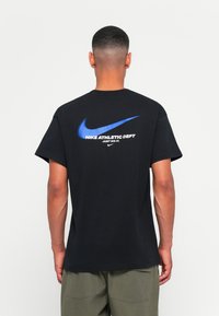 Fekete pamut póló, nagy kék Nike logóval és fehér szöveggel: "NIKE ATHLETIC DEPT JUST DO IT" a hátsó részén. Rövid ujjak, laza szabás.