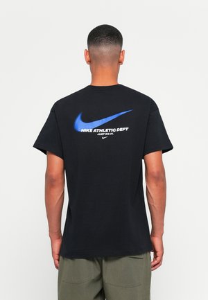 Melnas kokvilnas T-krekls ar lielu zilu Nike swoosh un baltu tekstu "NIKE ATHLETIC DEPT JUST DO IT" aizmugurē. Īsas piedurknes, brīvas piegriezuma.
