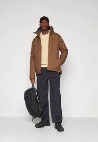 Hnědá zateplená bunda s kapucí, krémový pletený svetr, tmavé nákladní kalhoty a černý batoh. Celý outfit doplňují černé boty.