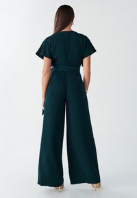 Tumši zaļa kombinezona ar īsām plandošām piedurknēm, jostu ap vidukli, platām pēdām un noslēptu aizmugurējo rāvējslēdzēju. Gluda, viegla auduma.