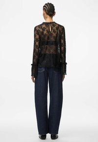 Crna bluza od čipke s dugim rukavima, s ključnim otvorom na leđima, naboranim manšetama i cvjetnim uzorcima. Uparen s širokim trapericama s nogavicama.