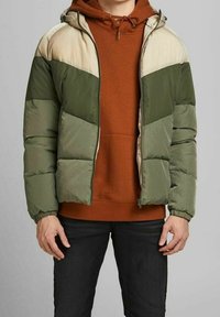 Padded jacket w kolorze ciemnozielonym i beżowym, z zapięciem na zamek, kapturem i elastycznymi mankietami. Noszona na rudym hoodie i ciemnych dżinsach.