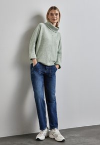 Ljusgrön, strukturerad, högkragad tröja i kombination med mörkblå jeans. Skorna är chunky vita sneakers med gråa detaljer.