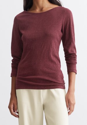 Vrouw draagt een bordeauxrode top met lange mouwen en een boothals, gecombineerd met lichtbeige broek, staand tegen een effen achtergrond.