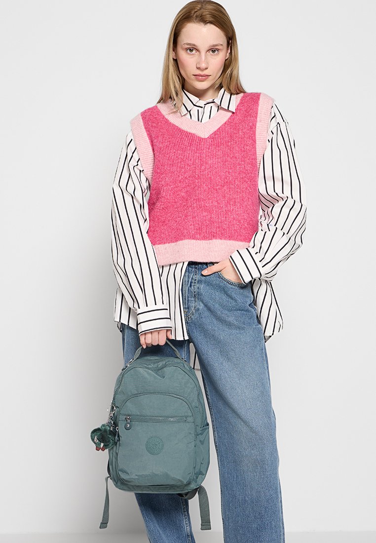 Joven mujer con un chaleco de punto rosa sobre una camiseta a rayas, jeans azules, sosteniendo una mochila color agua con un pequeño llavero de peluche.