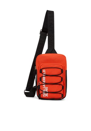 Kompaktna rdeča torbica čez ramo s črnim nastavljivim trakom, sprednjimi elastičnimi vrvicami in belim napisom "KARL LAGERFELD JEANS" na žepu.