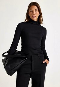 Musta ribbineuloksesta valmistettu poolopaita, yhdistetty räätälöityihin mustiin housuihin. Malli pitää suurta mustaa nahkalaukkua ja seisoo neutraalia taustaa vasten.