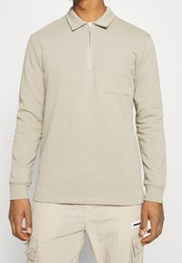 Homme portant un polo beige à manches longues avec une fermeture éclair et une poche poitrine, associé à un pantalon cargo beige clair avec des poches latérales.