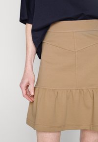 Gonna marrone chiaro realizzata in materiale morbido, con un rouche svasato all'orlo e dettagli cuciti, indossata con un top blu navy.