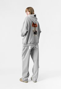 Sudadera gris de gran tamaño y pantalones de chándal a juego. La sudadera presenta un diseño de perro de dibujos animados en la parte posterior. Tejido suave, corte relajado, marca sutil.