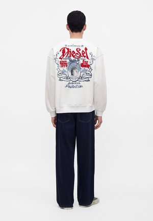 Homme de dos portant un sweat-shirt blanc avec un graphisme coloré et le texte "Diesel" et un pantalon large foncé sur fond uni.