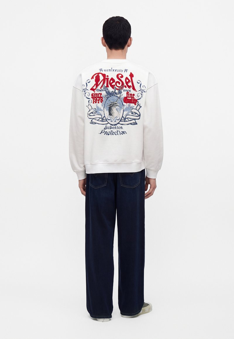 Homme de dos portant un sweat-shirt blanc avec un graphisme coloré et le texte "Diesel" et un pantalon large foncé sur fond uni.