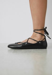 Sorte ballerinaer i læder med rundet tå, snørre-design ved anklen og metalkonter. Glat tekstur og minimalistisk silhuet.