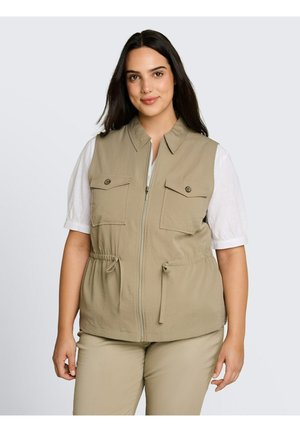 Frau mit langen dunklen Haaren, die eine beige Reißverschlussweste über einem weißen kurzärmeligen Hemd und passende beigen Hosen trägt, schaut nach vorne.