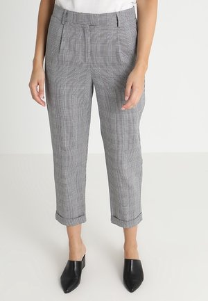 Personne portant un pantalon à carreaux gris longueur cheville et des mules noires à bout pointu, debout devant un fond clair uni.
