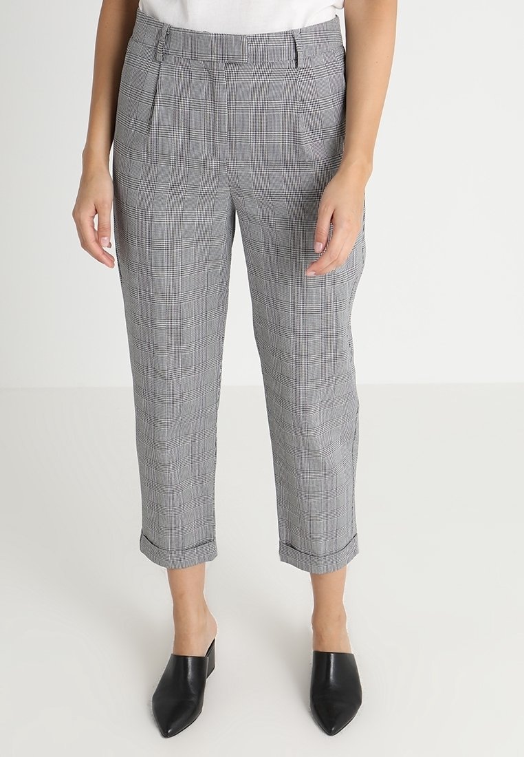 Personne portant un pantalon à carreaux gris longueur cheville et des mules noires à bout pointu, debout devant un fond clair uni.