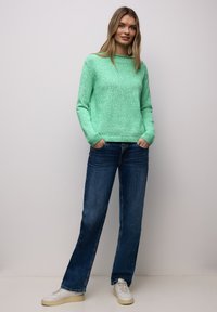 Jeune femme debout, mains dans les poches, portant un pull vert menthe, un jean bleu et des baskets blanches, devant un mur uni.